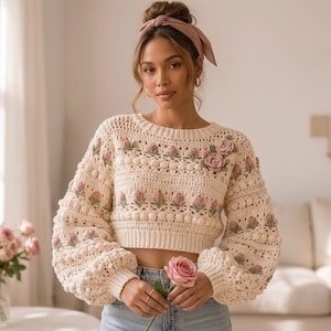 May include: A cream-coloured crochet sweater with pink floral detailing. The sweater has a round neckline, long, puffed sleeves, and a cropped style. The floral elements are clustered around the neckline and sleeves.