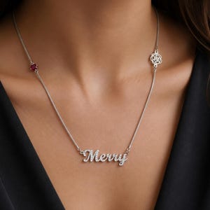 Könnte beinhalten: Eine silberne Halskette mit dem Namen "Merry" in funkelnden, klaren Kristallen. Die Halskette hat einen kleinen, runden roten Edelstein und ein dekoratives Blumendesign. Die zarte Kette hängt vor einem dunklen Hintergrund.