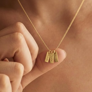 Può includere: Una collana color oro con tre piccoli ciondoli rettangolari verticali sospesi a una catenina sottile. Ogni ciondolo è inciso con un nome: Victoria, Taylor e Mason, scritto in corsivo sulla superficie metallica lucida.
