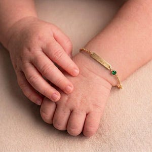 Può includere: Un bracciale a catena color oro con una targhetta identificativa rettangolare incisa con il nome Olivia e un piccolo simbolo a cuore, impreziosito da una singola pietra preziosa verde rotonda e sfaccettata incastonata in una montatura dorata.
