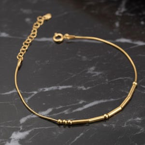May include: A delicate gold bracelet featuring a fine chain and a series of small, cylindrical and spherical beads. The bracelet has an adjustable chain and a clasp closure. The jewellery is displayed on a dark, marbled surface.