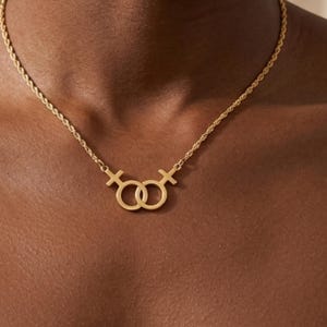 Op de afbeelding: Goudkleurige ketting met een hanger met twee in elkaar grijpende vrouwelijke symbolen. De ketting heeft een delicaat, gedraaid touwontwerp. De ketting wordt gedragen op een donkere huidskleur, waardoor een contrast ontstaat dat de sieraden benadrukt.