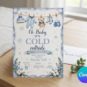 May include: A baby shower invitation card featuring a blue gingham border and a clothesline with a winter hat, onesie, teddy bear, socks, and mittens. The text reads: Oh Baby, IT'S COLD outside, PLEASE JOIN US FOR A BABY SHOWER, Amanda Smith, January 18th, 2026.