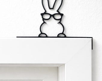 May include: Black metal bunny silhouette wearing glasses, designed to sit on a door frame. The bunny has long ears and a round body, adding a touch of whimsy to home decor. The glasses are round and the bunny is perched on a white door frame.