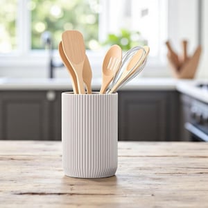 Könnte beinhalten: Ein weißer, geriffelter Keramik-Utensilienhalter auf einem Holztisch, gefüllt mit Holzlöffeln und einem Schneebesen. Das Küchenutensil steht vor einem verschwommenen Küchenhintergrund mit einem Fenster.