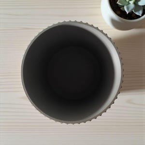 May include: An overhead view of a gray, round planter with a textured rim. The interior is dark gray. A small white ceramic pot with a succulent plant sits nearby on a light wood surface. The planter is empty.