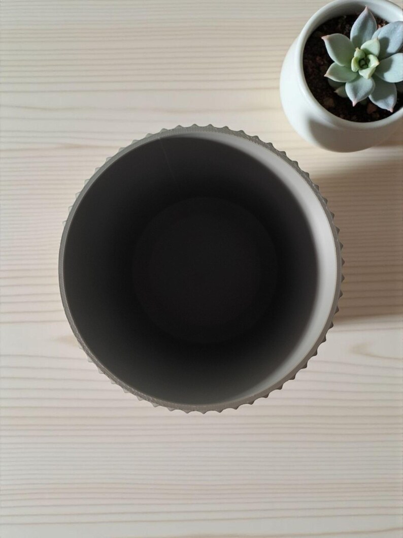May include: An overhead view of a gray, round planter with a textured rim. The interior is dark gray. A small white ceramic pot with a succulent plant sits nearby on a light wood surface. The planter is empty.