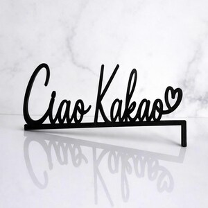 May include: Black cursive sign that reads "Ciao Kakao" with a heart symbol. The sign is made of a solid material and has a rectangular base for stability. The sign is displayed on a white surface.