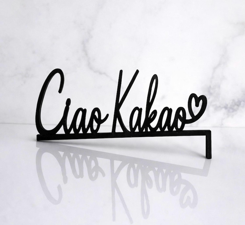 May include: Black cursive sign that reads "Ciao Kakao" with a heart symbol. The sign is made of a solid material and has a rectangular base for stability. The sign is displayed on a white surface.