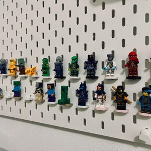May include: A white pegboard displaying a collection of Lego minifigures. The figures are arranged on small white shelves, showcasing a variety of characters in different colors and designs. The pegboard is mounted on a white wall.