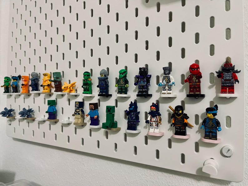 May include: A white pegboard displaying a collection of Lego minifigures. The figures are arranged on small white shelves, showcasing a variety of characters in different colors and designs. The pegboard is mounted on a white wall.