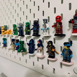 May include: A collection of colorful Lego minifigures displayed on a white pegboard. The figures feature various designs and colors, including red, green, blue, and black. They are mounted on small, round, pink and black platforms.