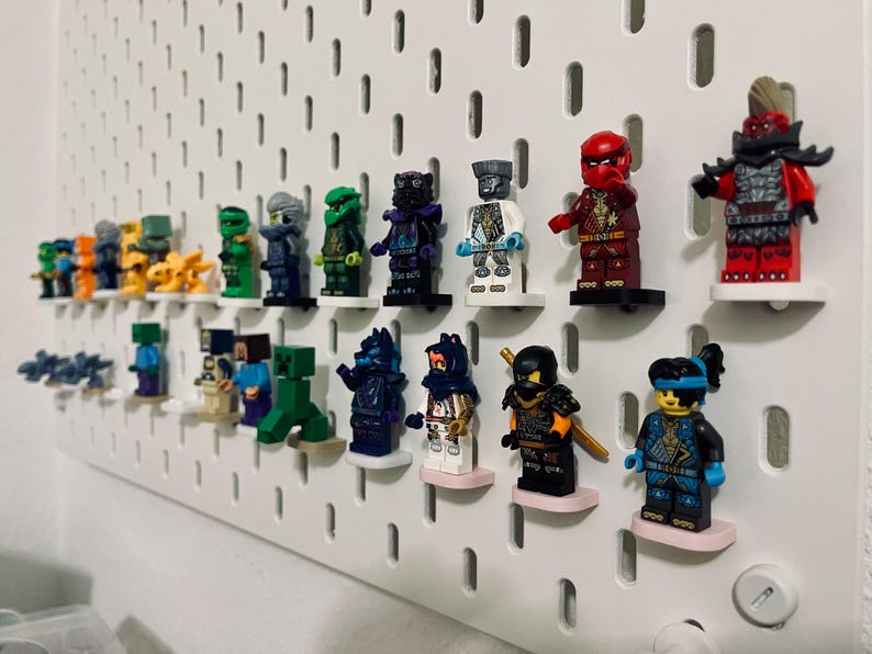 May include: A collection of colorful Lego minifigures displayed on a white pegboard. The figures feature various designs and colors, including red, green, blue, and black. They are mounted on small, round, pink and black platforms.