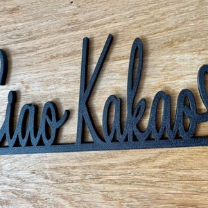 May include: Black metal sign with the words "Ciao Kakao" in a cursive font, with a heart replacing the letter "o" at the end. The sign is designed to be placed on a surface.