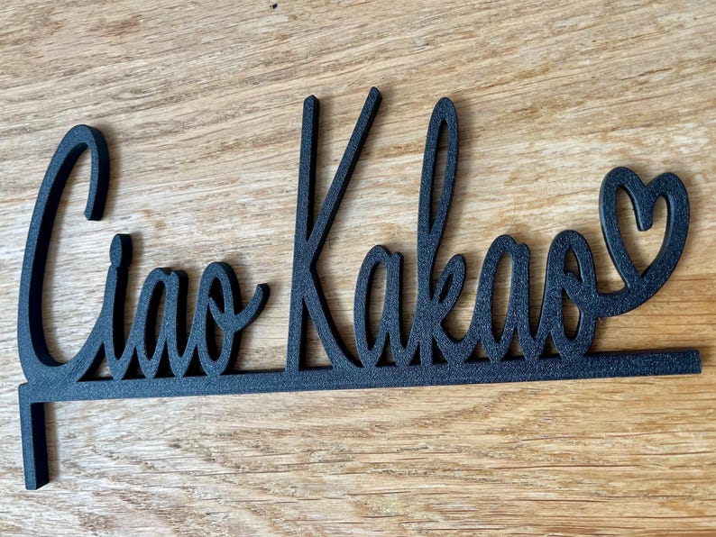May include: Black metal sign with the words "Ciao Kakao" in a cursive font, with a heart replacing the letter "o" at the end. The sign is designed to be placed on a surface.