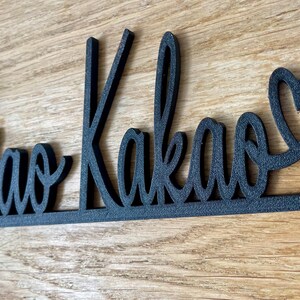 May include: Black wooden sign with the words "Ciao Kakao" in a cursive font, with a heart replacing the letter "o" at the end. The sign is on a wooden surface.