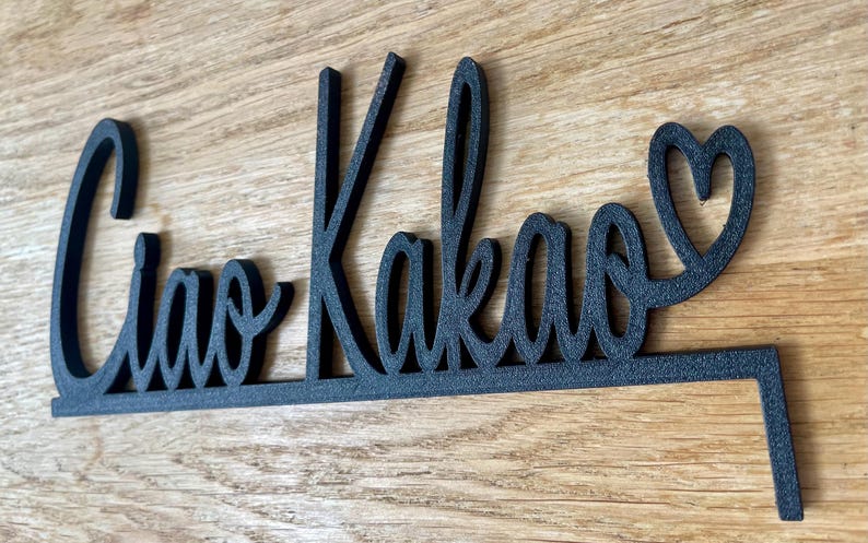 May include: Black wooden sign with the words "Ciao Kakao" in a cursive font, with a heart replacing the letter "o" at the end. The sign is on a wooden surface.