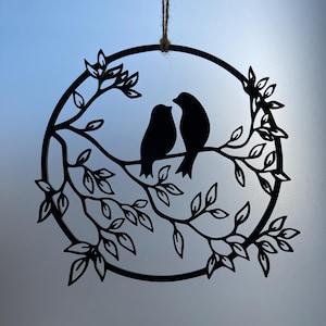 Op de afbeelding: Zwarte metalen wandkunst met twee vogels op een tak in een cirkelvormige lijst. Het ontwerp bevat bladeren en takken, opgehangen aan een natuurlijke touw. Geschikt voor interieurdecoratie.