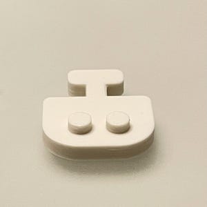 May include: A close-up of a white, plastic boat-shaped object. The object has a rectangular base with two small, round protrusions and a T-shaped handle. The background is a solid, light beige color.