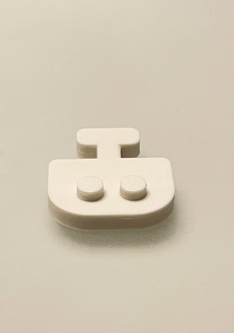 May include: A close-up of a white, plastic boat-shaped object. The object has a rectangular base with two small, round protrusions and a T-shaped handle. The background is a solid, light beige color.