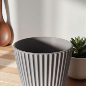 May include: A gray, ribbed ceramic planter with a wide opening. The planter has vertical grooves and a slightly textured surface. A small white pot with a green succulent is to the right. A wooden spoon is in the background.