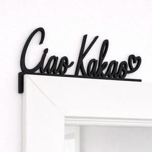 May include: Black wooden sign with the words "Ciao Kakao" in cursive script, with a heart replacing the second "o". The sign is mounted on a white door frame.