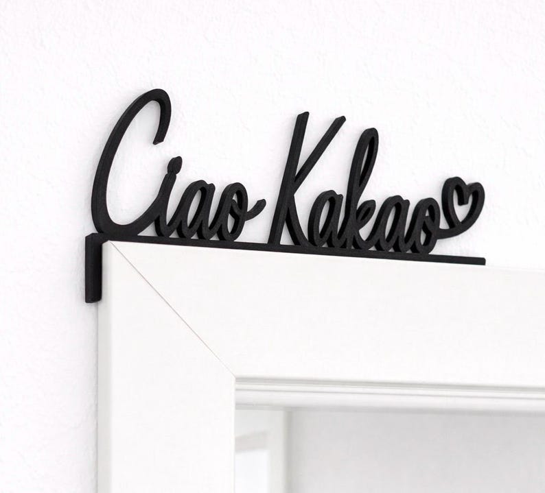 May include: Black wooden sign with the words "Ciao Kakao" in cursive script, with a heart replacing the second "o". The sign is mounted on a white door frame.