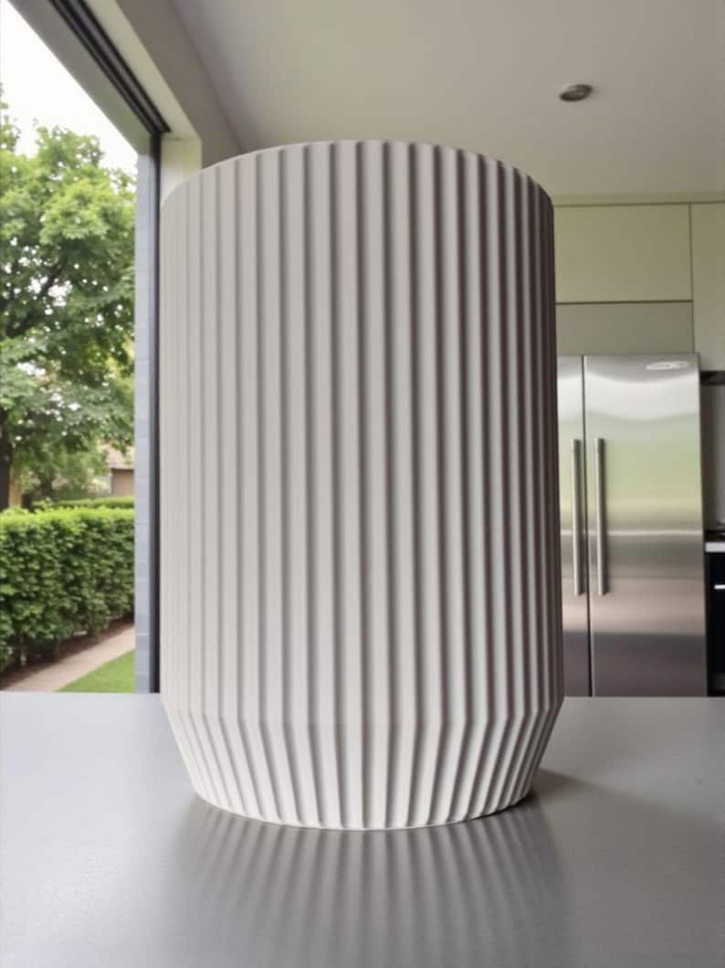 May include: A tall, cylindrical, off-white vase with vertical ribbed detailing. The vase has a slightly flared base and stands on a gray surface. The background includes a window with a view of greenery and a modern kitchen.