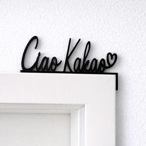 May include: Black wooden sign with the words "Ciao Kakao" in a cursive font, with a heart symbol at the end. The sign is mounted on a white door frame, creating a decorative accent.