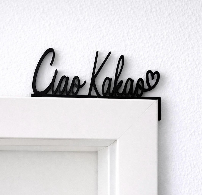 May include: Black wooden sign with the words "Ciao Kakao" in a cursive font, with a heart symbol at the end. The sign is mounted on a white door frame, creating a decorative accent.