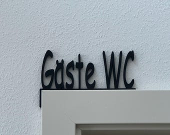 May include: A black, decorative door sign featuring the text "Gäste WC" in a stylized, cursive font. The sign is designed with an L-shaped bracket that rests securely on the top corner of a door frame, providing a clear label for a guest restroom.