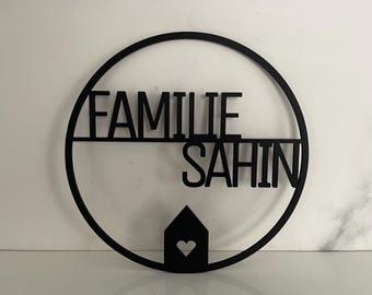 Könnte beinhalten: Schwarzes Metall-Familienschild mit dem Wort "FAMILIE" über "SAHIN" in einem kreisförmigen Rahmen. Eine Haussilhouette mit einem Herz-Ausschnitt ist unter den Namen zentriert. Für die Inneneinrichtung.