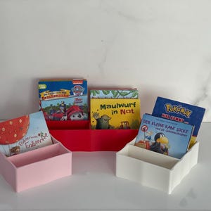 Peut inclure: Trois petits organiseurs de livres roses, rouges et blancs, chacun contenant des livres pour enfants. Les livres ont des couvertures colorées avec des titres comme "Pat' Patrouille" et "Pokemon". Les organiseurs sont disposés sur une surface blanche.