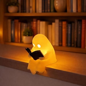 Könnte beinhalten: Eine leuchtend gelbe, geisterförmige Lampe, die ein schwarzes Buch liest und auf einem Holzabsatz sitzt. Die Lampe hat zwei helle, augenförmige Lichter. Im Hintergrund ein Bücherregal voller Bücher.
