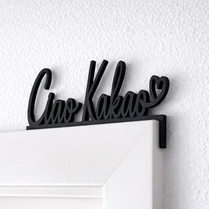 May include: Black wooden sign with the words "Ciao Kakao" in cursive script, with a heart symbol at the end. The sign is designed to hang over a door frame. The door frame is white, and the wall is white.