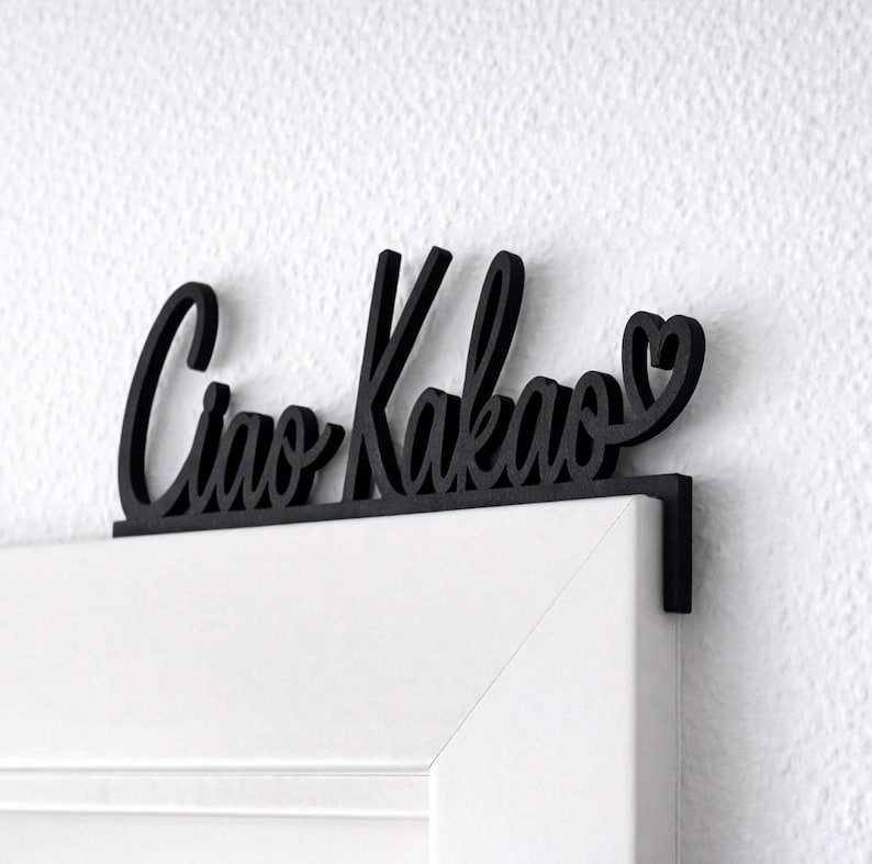 May include: Black wooden sign with the words "Ciao Kakao" in cursive script, with a heart symbol at the end. The sign is designed to hang over a door frame. The door frame is white, and the wall is white.