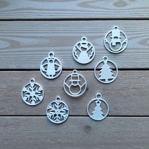 Könnte beinhalten: Eine Sammlung weißer Weihnachtsschmuckstücke mit verschiedenen Designs, darunter Engel, Schneeflocken und Weihnachtsbäume. Jedes Ornament ist kreisförmig mit einer kleinen Schlaufe zum Aufhängen. Die Ornamente sind auf einer Holzoberfläche angeordnet.