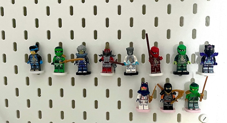 May include: A collection of colorful Lego Ninjago minifigures, each with unique armor and accessories. The figures are displayed on a white pegboard, showcasing a variety of ninja characters in vibrant colors like red, green, and blue.