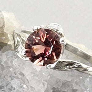 May include: A silver ring featuring a large, round, faceted, reddish-brown gemstone. The ring's band has a delicate, twisted design, and the gemstone is held in place by four prongs. The ring is set against a textured, light-colored background.
