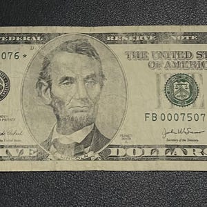 May include: A five-dollar bill featuring a portrait of Abraham Lincoln. The bill displays the text "FIVE DOLLARS" and "THE UNITED STATES OF AMERICA". Serial number FB 00075076* is visible.