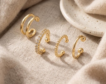 May include: A set of four gold-toned ear cuffs. The first is a double-banded smooth cuff, the second features a row of small clear stones, the third has a twisted rope texture, and the fourth is a simple smooth band with small ball ends.