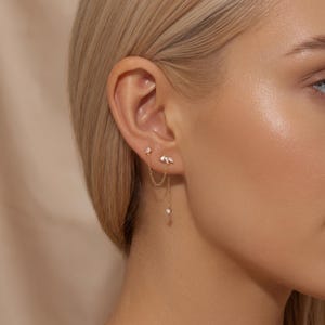 Può includere: Un orecchino a catena dorata con due perni collegati da una sottile catenina. Il perno superiore è una pietra a forma di goccia, mentre quello inferiore è composto da tre piccole pietre taglio marquise con un minuscolo cristallo pendente.