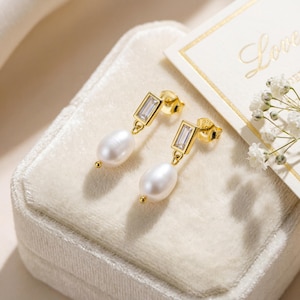 May include: A pair of gold-colored earrings with a rectangular, clear stone and a dangling white pearl. The earrings are displayed on a white velvet surface, with a small white flower arrangement and a card that says "Love" in the background.
