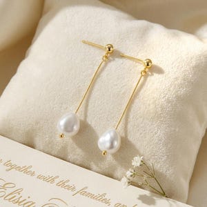 May include: A pair of elegant gold-toned drop earrings featuring lustrous, white, teardrop-shaped pearls. The earrings have a delicate, linear design with a small gold ball at the top and bottom of each pearl. The earrings are displayed on a cream-colored surface.