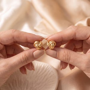 May include: A pair of gold-toned stud earrings, each featuring a three-dimensional knot design with a smooth, polished finish. The earrings are spherical in shape, composed of intertwined metal loops that create a continuous, rounded knot pattern.