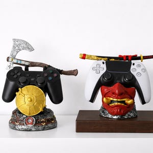 Może przedstawiać: Dwa stojaki na kontrolery do gier. Jeden ma czarny kontroler ze srebrną siekierą i złotym dyskiem. Drugi ma biało-czarny kontroler z czerwoną maską i kataną. Oba są dekoracyjne.