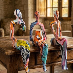 May include: Three abstract sculptures of seated figures with colourful, swirled paint designs. The figures are perched on a wooden table, each with a unique pose and colour palette, including orange, blue, and yellow.