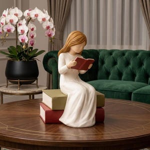 Könnte beinhalten: Keramikfigur einer Person, die ein Buch liest und auf einem Stapel Bücher sitzt. Die Figur hat lange, hellbraune Haare und trägt ein weißes Kleid. Die Bücher sind rot und grün. Eine Topf-Orchidee und ein grünes Sofa befinden sich im Hintergrund.