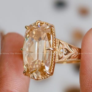 May include: A gold ring featuring a large, elongated, champagne-colored gemstone held by multiple prongs. The band is crafted with intricate, engraved floral-like patterns and includes a small, clear accent stone on the side of the setting.