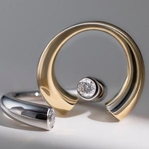 May include: A two-tone open ring featuring a polished gold-colored curved band and a silver-colored curved band. Each end of the bands is set with a single round brilliant-cut white diamond, creating a modern bypass design with contrasting metal tones.
